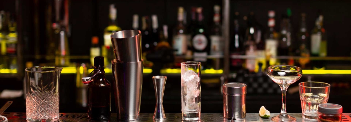 Does your business require a Liquor Licence and how do you apply for one? Picture of a bar with cocktail shakers and ornate glasses lined up in a row. Rows of alcoholic drink bottles lined up in the background.