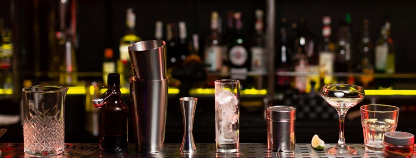 Does your business require a Liquor Licence and how do you apply for one? Picture of a bar with cocktail shakers and ornate glasses lined up in a row. Rows of alcoholic drink bottles lined up in the background.